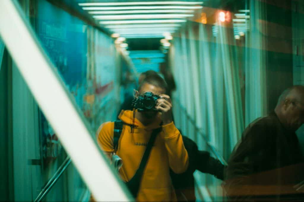 A gay guy in a yellow hoodie holds a camera, reflecting in a glass surface at the airport, with a blurred background of other people.