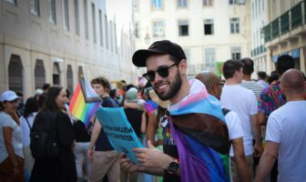 A man beams with pride at a parade, draped in a rainbow flag cape and holding a sign—a celebration of identity and the benefits of gay travel, highlighting the vibrant connections that unite LGBTQ+ travelers worldwide.
