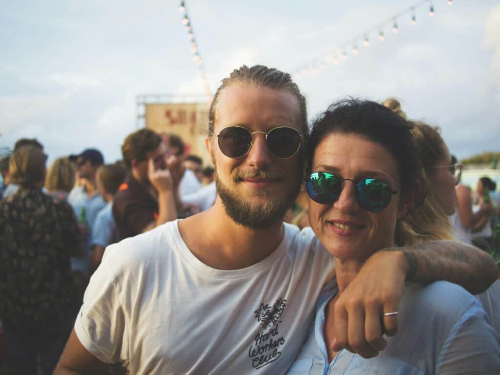 Mother and gay son smiling at a festive outdoor event, surrounded by a crowd. They wear sunglasses.
