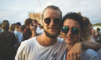 Mother and gay son smiling at a festive outdoor event, surrounded by a crowd. They wear sunglasses.
