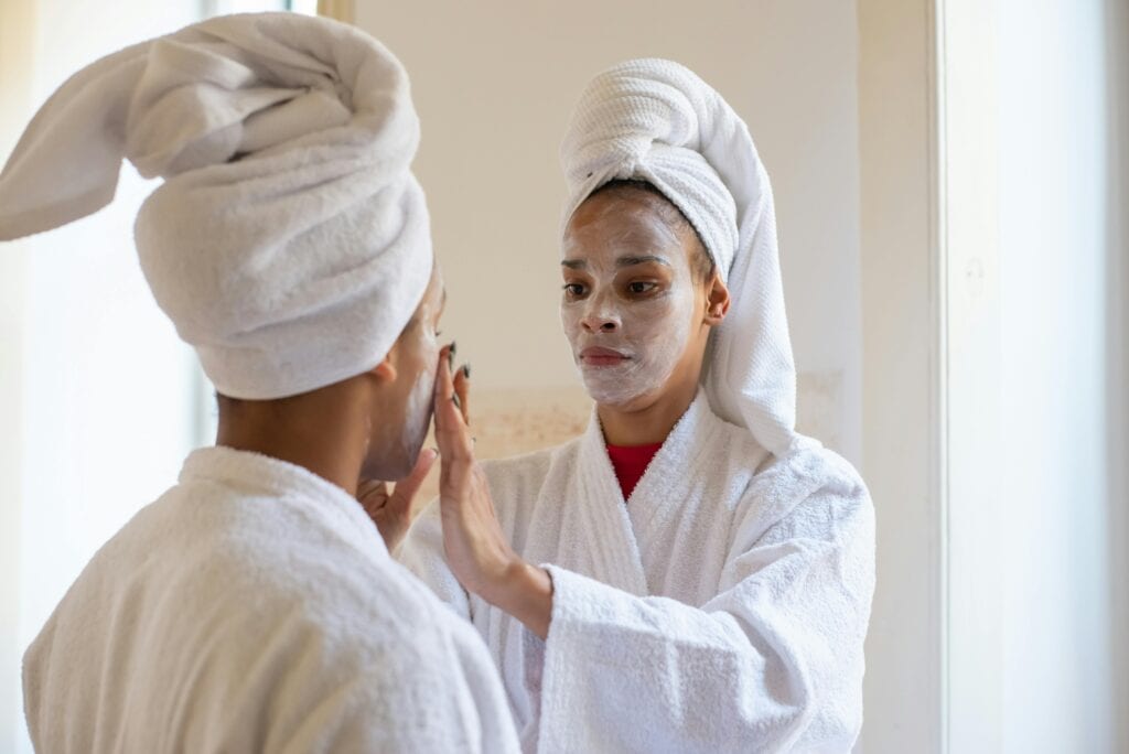 A mother and son in white robes and towels apply face masks in front of a mirror at the spa, celebrating with Mother’s Day travel gifts.