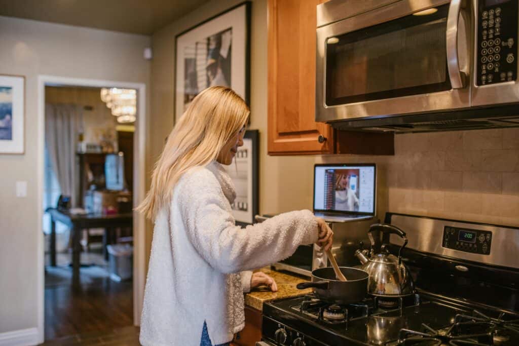 A mother with long blonde hair stirs a pot on the stove while following a virtual cooking class on her laptop, enjoying Mother’s Day travel gifts.