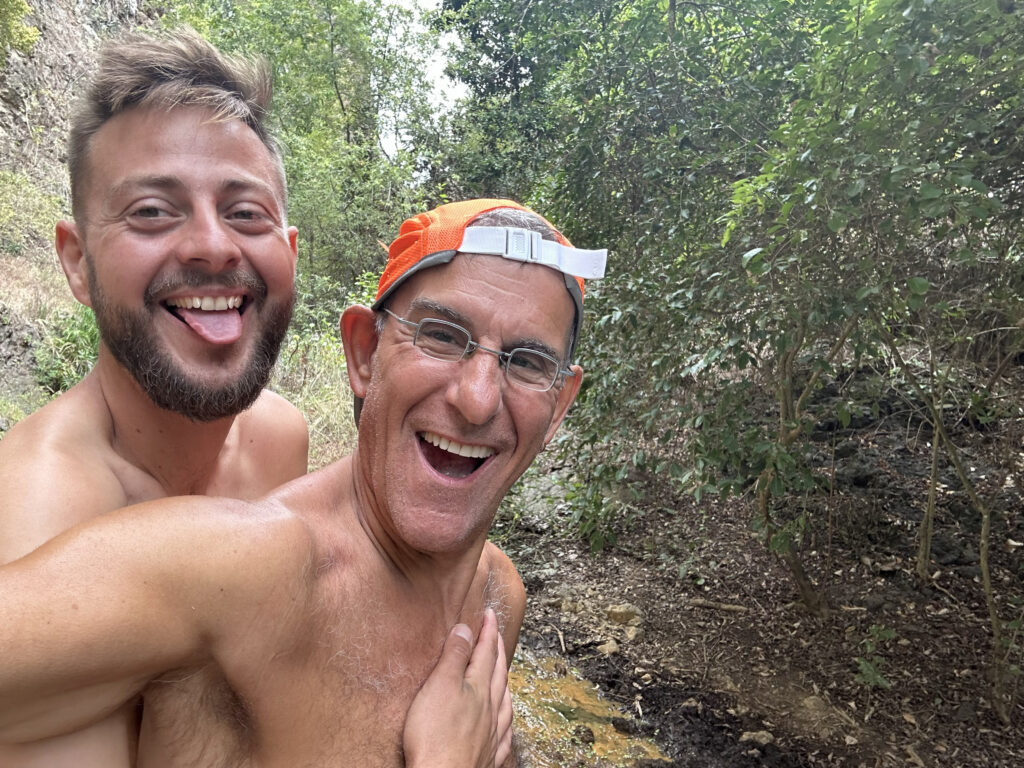 Two smiling men, one in a cap, enjoying nature on a lush green pathway during their naked vacation for men with Everything To Sea.