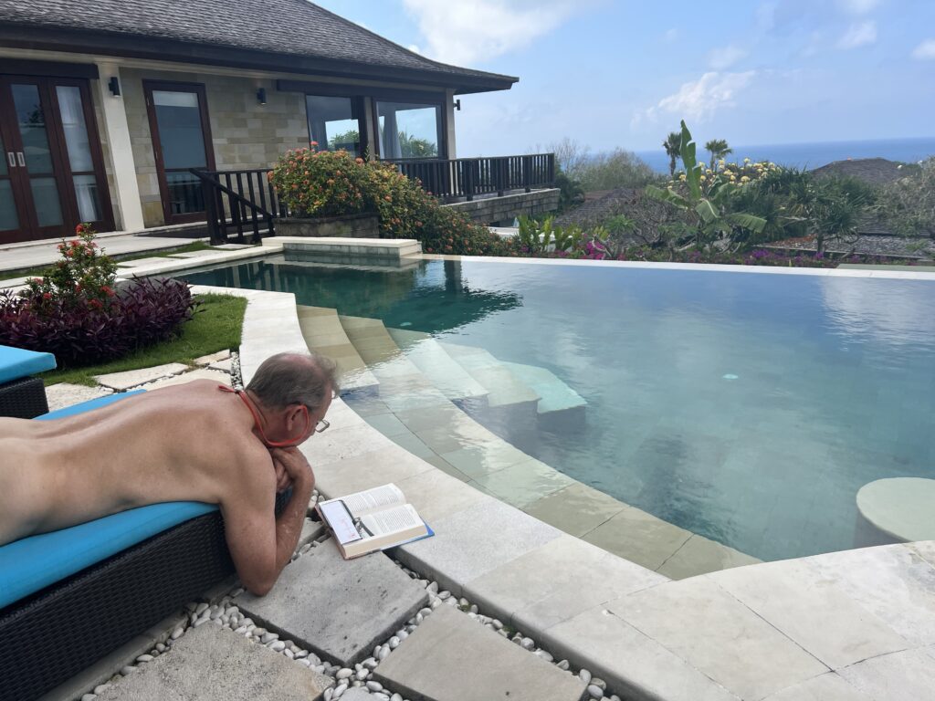 A man enjoying a naked vacation for men with Everything To Sea, relaxing by an infinity pool, reading a book, and surrounded by lush tropical plants.