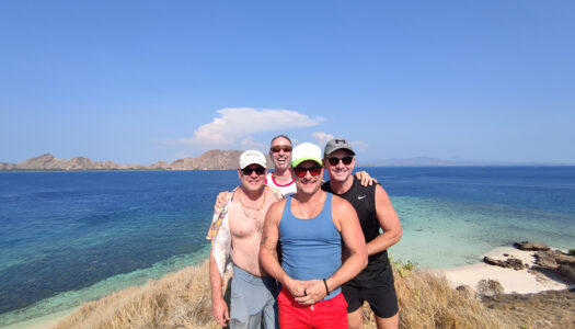 Group of four men smiling near the coast with a blue ocean and green islands in the background, enjoying a naked vacation for men on a trip with Everything To Sea.
