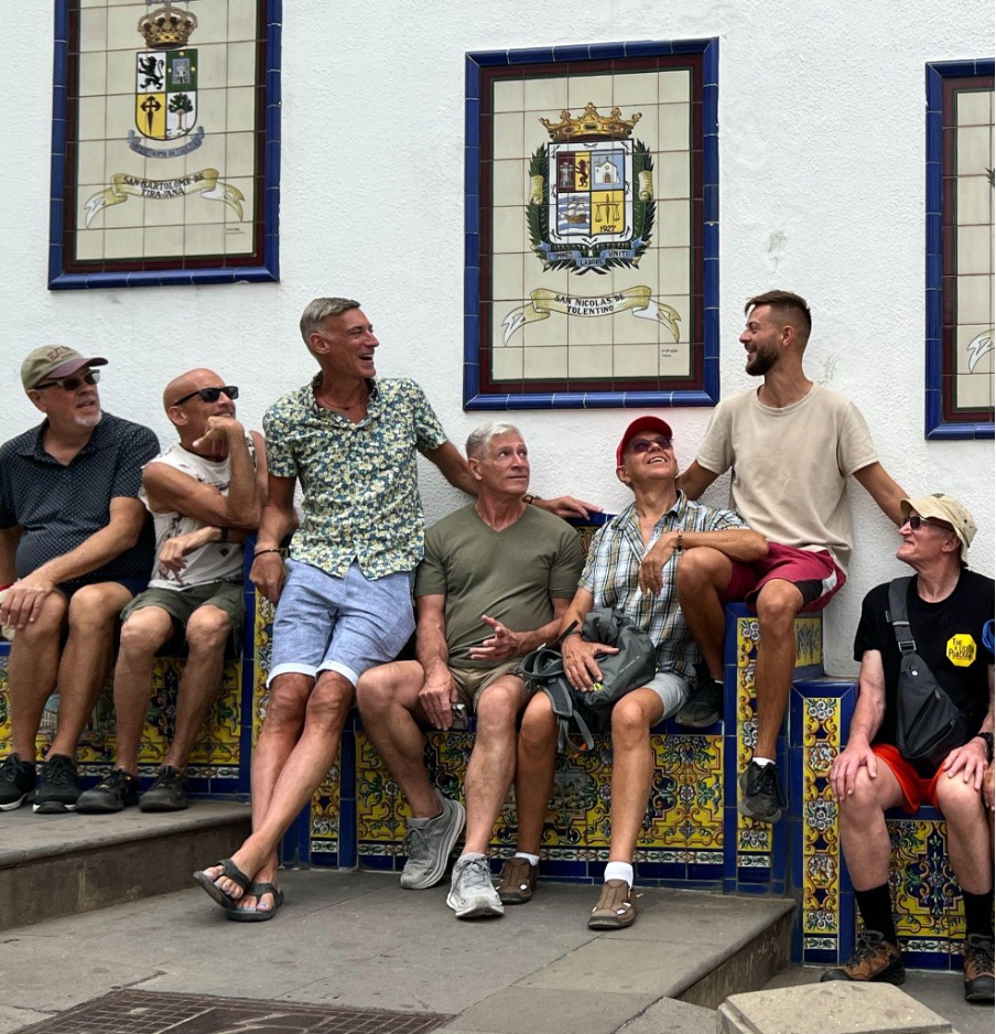 Seven men sitting on colorful tiles, laughing and interacting with each other during a naked vacation for men with Everything To Sea.