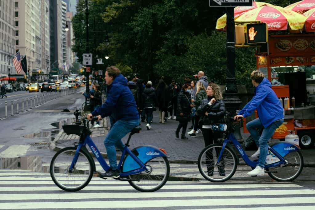 Two riders on blue Citi Bikes, part of a popular bike-sharing program for travelers, crossing a busy street in New York City. People walk nearby.