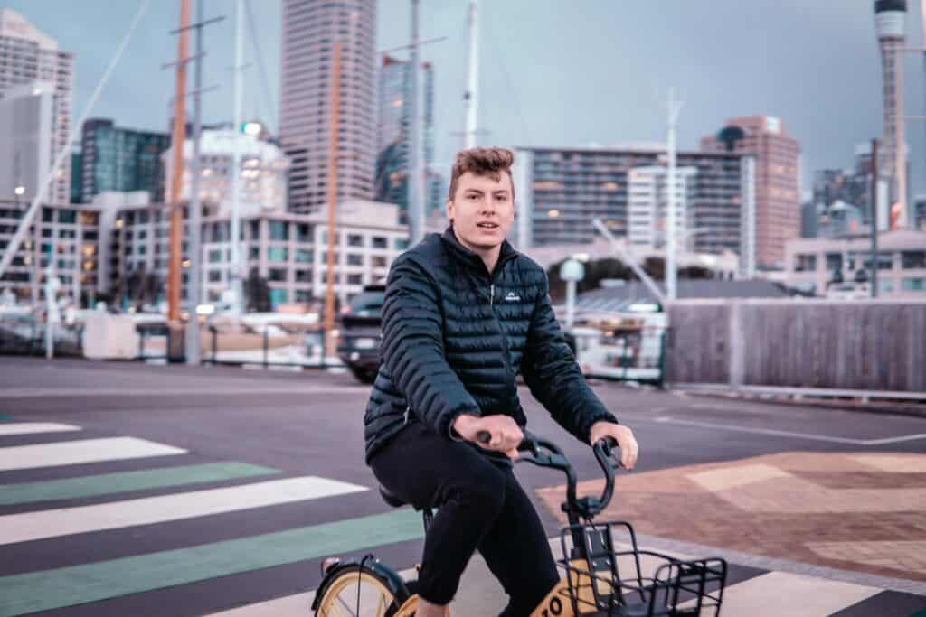 A person in a black jacket riding a bike near a marina with buildings in the background, showcasing the convenience of bike sharing for travelers exploring the area.