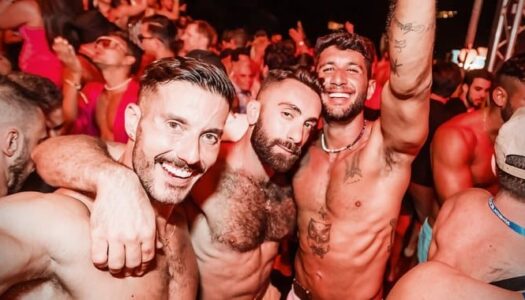 Shirtless men dancing and smiling at a packed gay circuit party, capturing the energy of gay travel peak sluttiness nightlife.