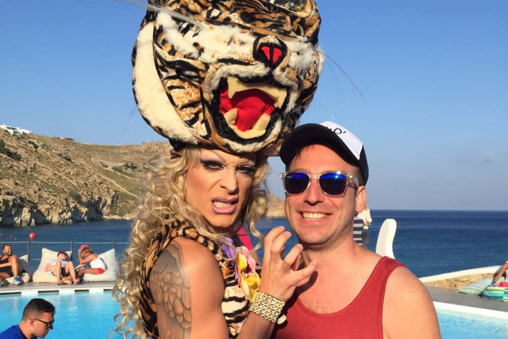 Drag queen in an elaborate tiger headdress posing with a smiling traveler at JackieO’s Mykonos gay bar by the seaside pool.