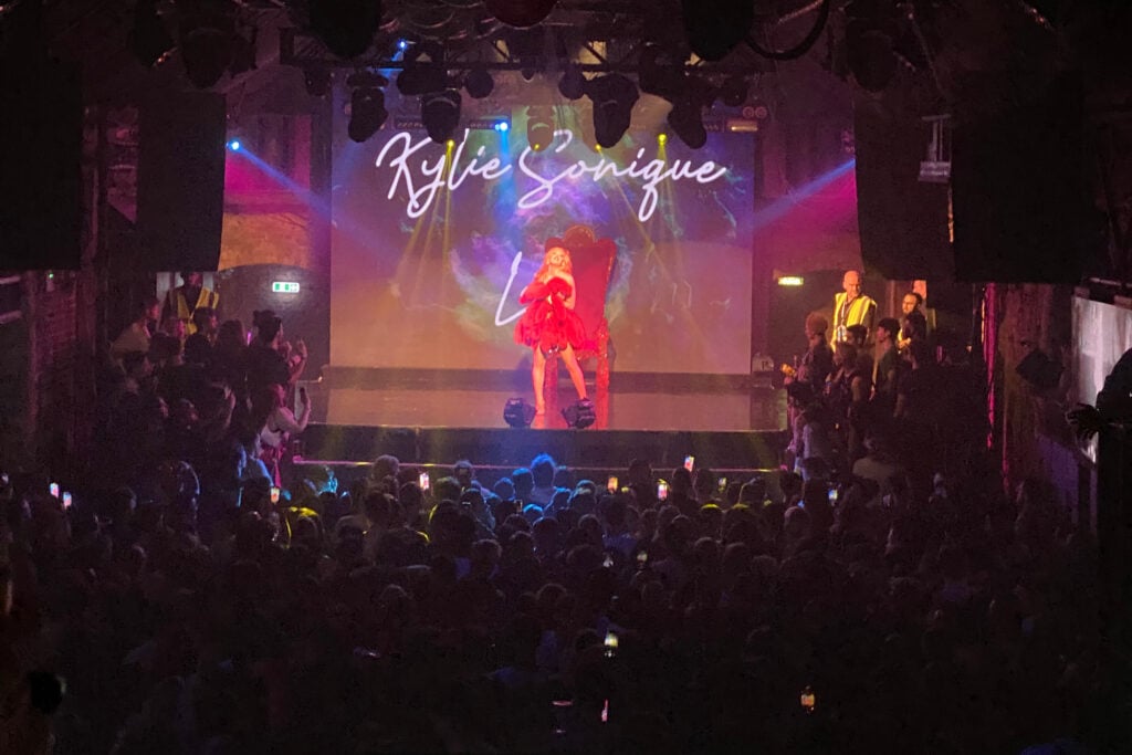 Kylie Sonique, a drag queen, performing on stage under bright lights at Heaven gay bar in London, with a large cheering audience.