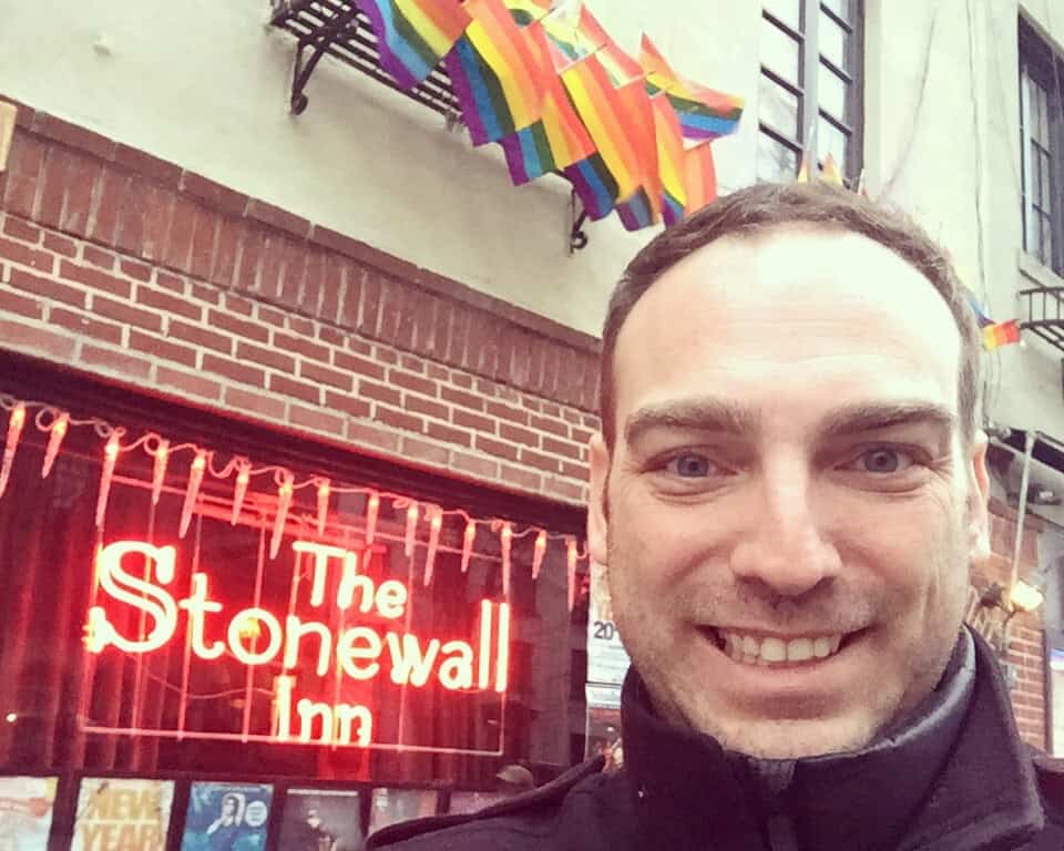 Handsome traveler smiling outside The Stonewall Inn gay bar in New York City, with rainbow flags and neon sign in the background.