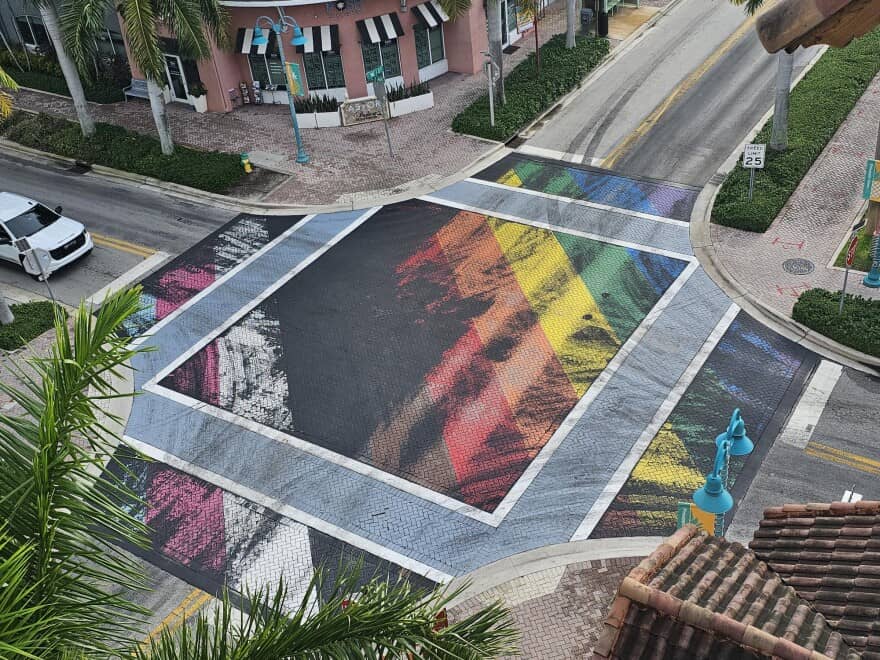 Aerial view of rainbow crosswalks in Florida controversy, showing a colorful Pride flag design painted across an intersection with palm trees and shops nearby.