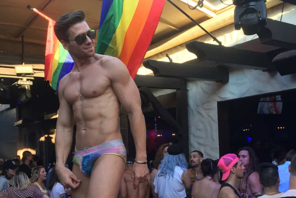 Shirtless go-go dancer in briefs performing at The Abbey gay bar in Los Angeles, surrounded by rainbow flags and partygoers.