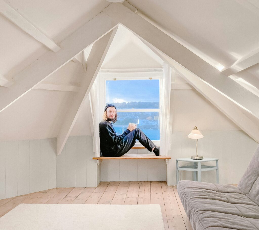 Man sitting in a cozy Airbnb loft window overlooking water, showing budget-friendly stays as part of cheap gay travel hacks.