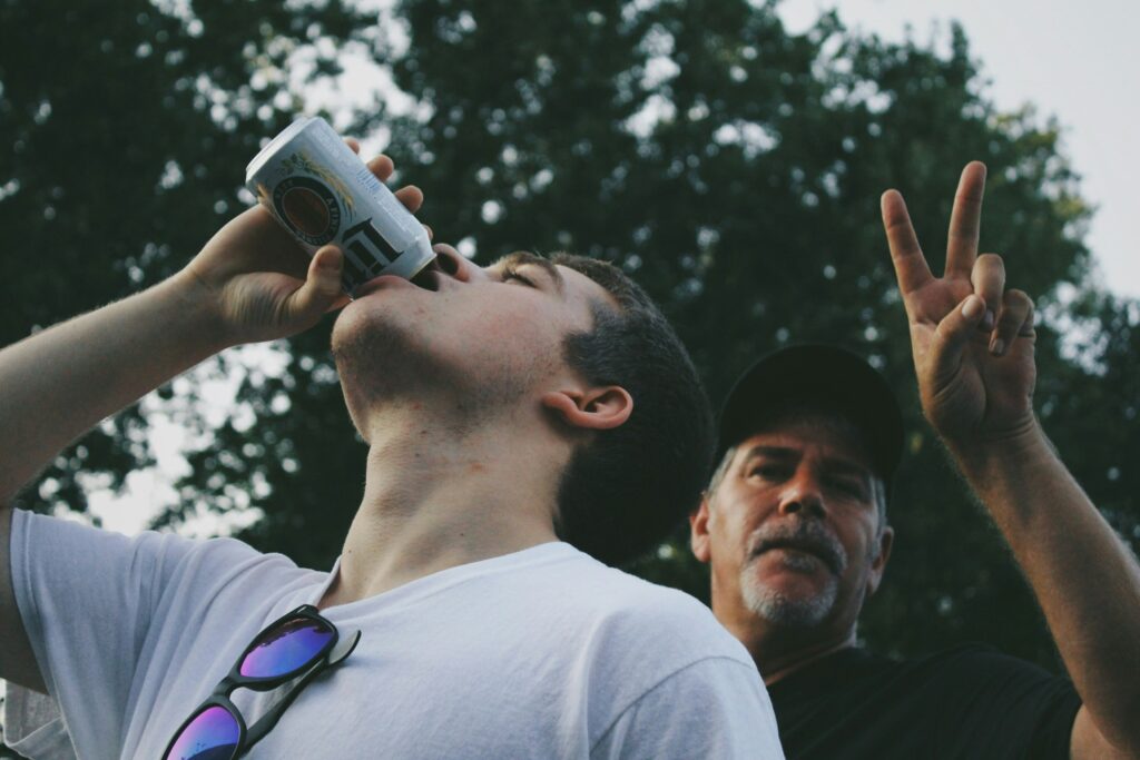 Young man drinking a beer outdoors with a friend flashing a peace sign, a casual moment of cheap gay travel nightlife.