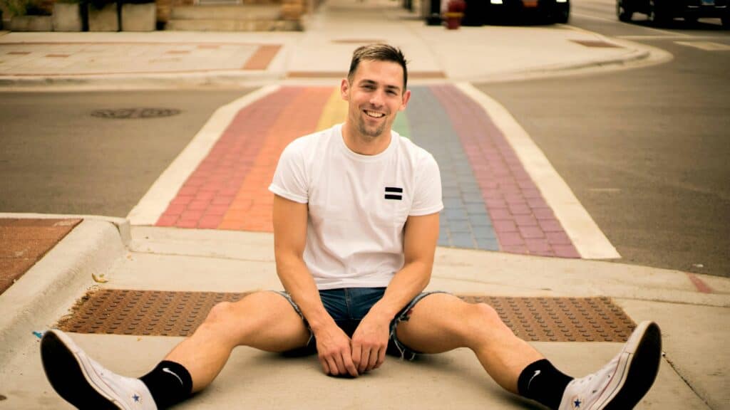 Young man sitting on rainbow crosswalk during Florida controversy, smiling in front of a faded Pride flag design painted on the street.