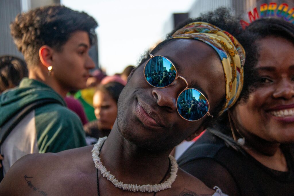 Pride attendee in sunglasses and necklace smiling at the camera in a vibrant crowd, a joyful cheap gay travel experience.