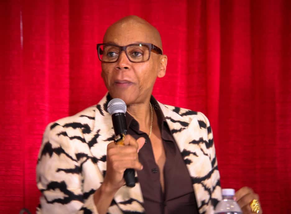 RuPaul speaking into a microphone in front of a red curtain, wearing glasses and a bold patterned blazer.