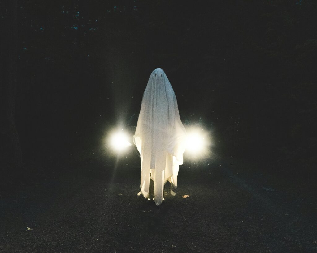 A person dressed as a ghost in a white sheet stands in front of a car with headlights on at night, symbolizing visibility and awareness for Halloween car safety tips.