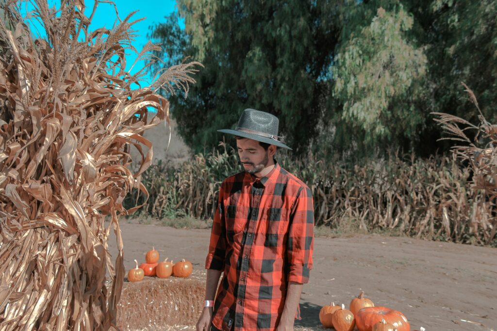 A man in a red flannel shirt and wide-brimmed hat stands in a pumpkin patch surrounded by dried corn stalks at a fall festival.