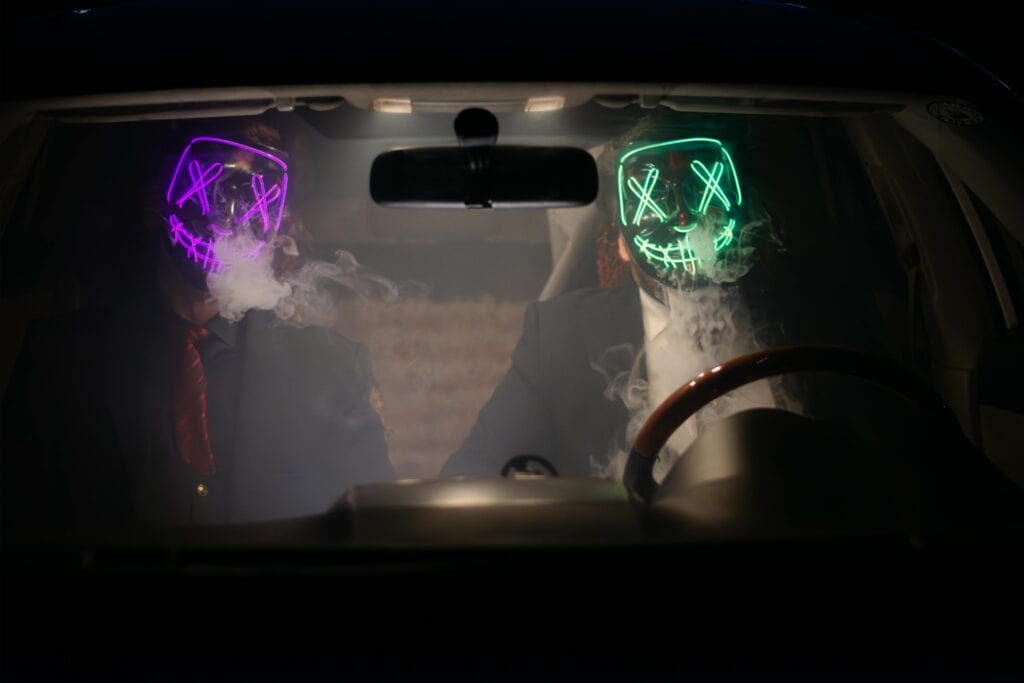 Two people wearing glowing neon masks sit inside a smoky car at night, illustrating the importance of Halloween car safety tips about avoiding distractions and impaired driving.