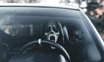 A driver wearing a Scream mask grips the steering wheel inside a car, emphasizing Halloween car safety tips and the need for focused, cautious driving on spooky nights.