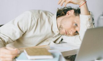A tired man rests his head on his arm beside a laptop and notebooks, showing the struggle of low energy and how to fight fall fatigue at work.