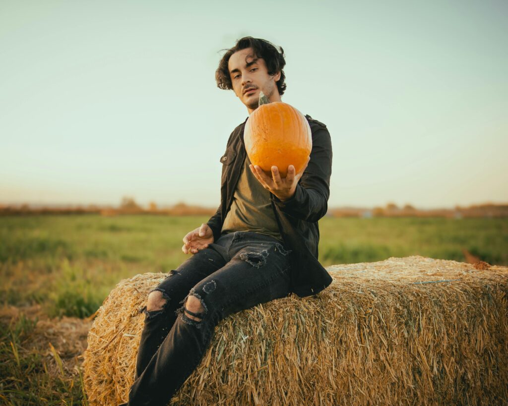 A man sitting on a hay bale in a field holds a pumpkin at sunset during a fall festival.