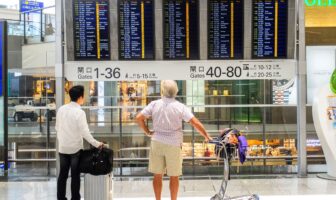 Two travelers checking airport departure boards while planning how to find cheap flights.