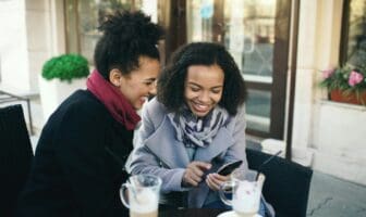 Two women sharing a smartphone at a café, illustrating lesbian dating community apps.