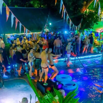 A lively nighttime pool party at Bourbon Street Pub Garden Bar in Key West, Florida, featuring a vibrant gay social scene. The tropical atmosphere is adorned with colorful pennant banners, festive lighting, and palm trees, with attendees in swimwear mingling around two pools. The energy is electric, offering a unique and inclusive experience for those exploring the best gay-friendly activities in Key West.